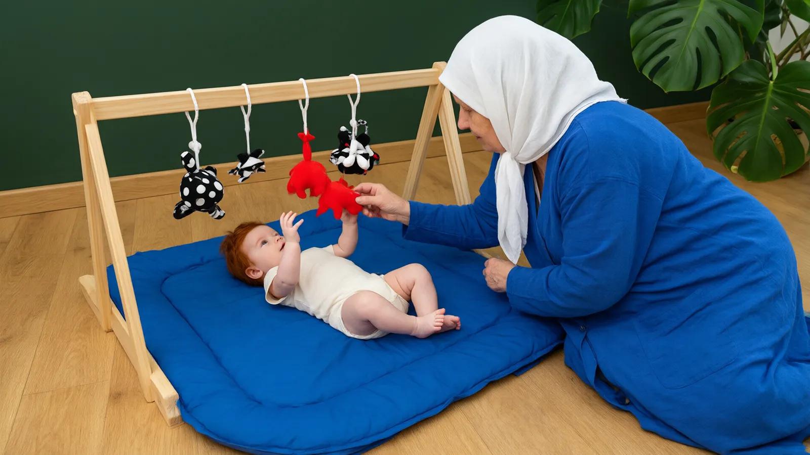 مادربزرگی با روسری سفید کنار نوزاد سهماهه روی تشک آبی زیر آویز اسباببازی در اتاقی با دیوار سبز تیره است