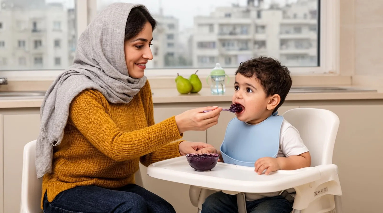 مادر ایرانی با شال روشن در آشپزخانهٔ اپن آپارتمان مدرن، کودک ۱۸ ماهه‌اش را روی صندلی غذا نشانده و با قاشق پورهٔ آلو می‌دهد؛ لیوان آب کودک و چند گلابی روی میز است